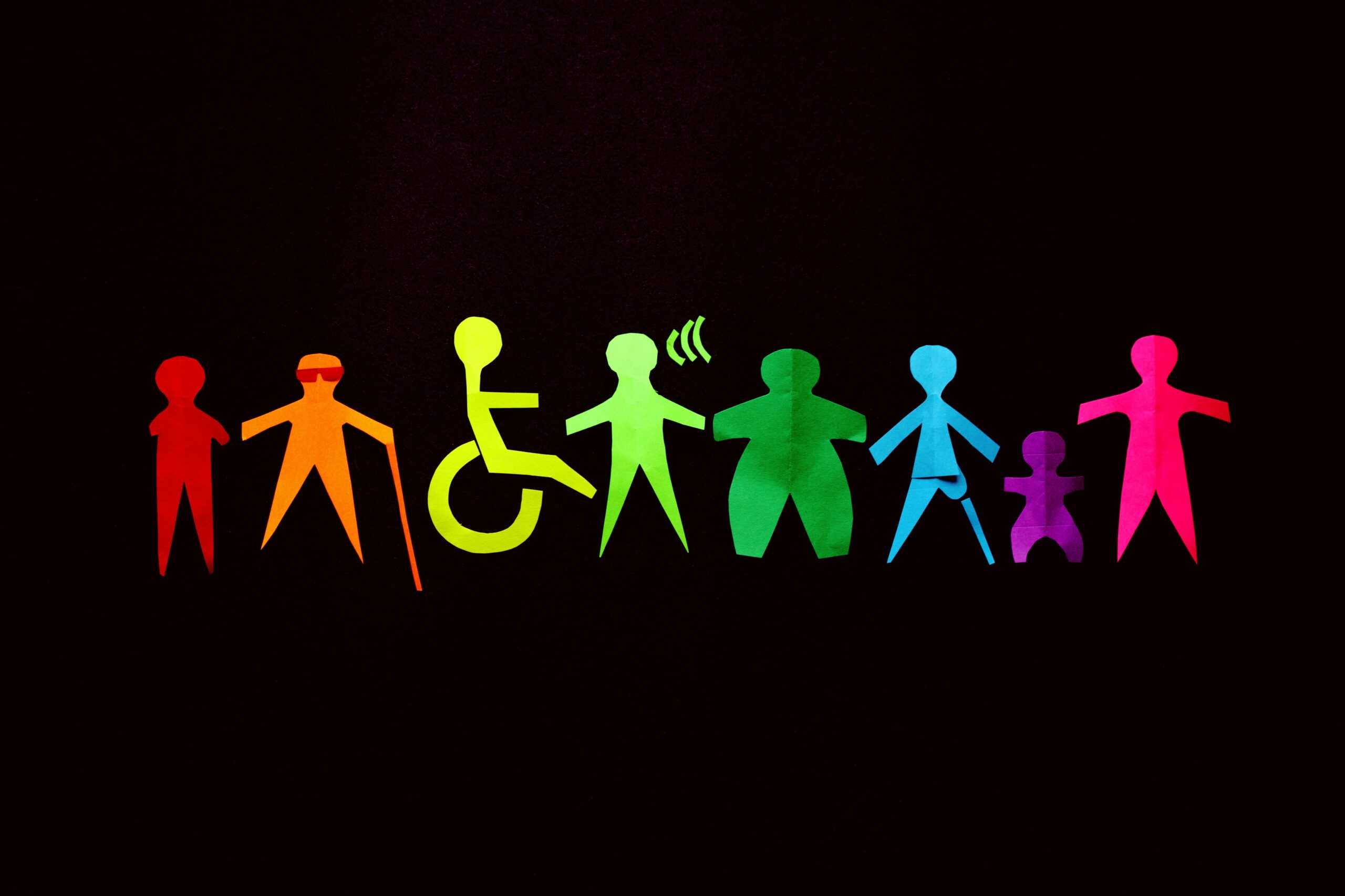 A group of rainbow colored stick figures with various disabilities are situated in a line. The stick figures show a blind person with a white cane, a person using a wheelchair, a person with dwarfism, people with limb differences and a Deaf person, all with varying body types. Each disability is valid and worthy of accessible accommodations.