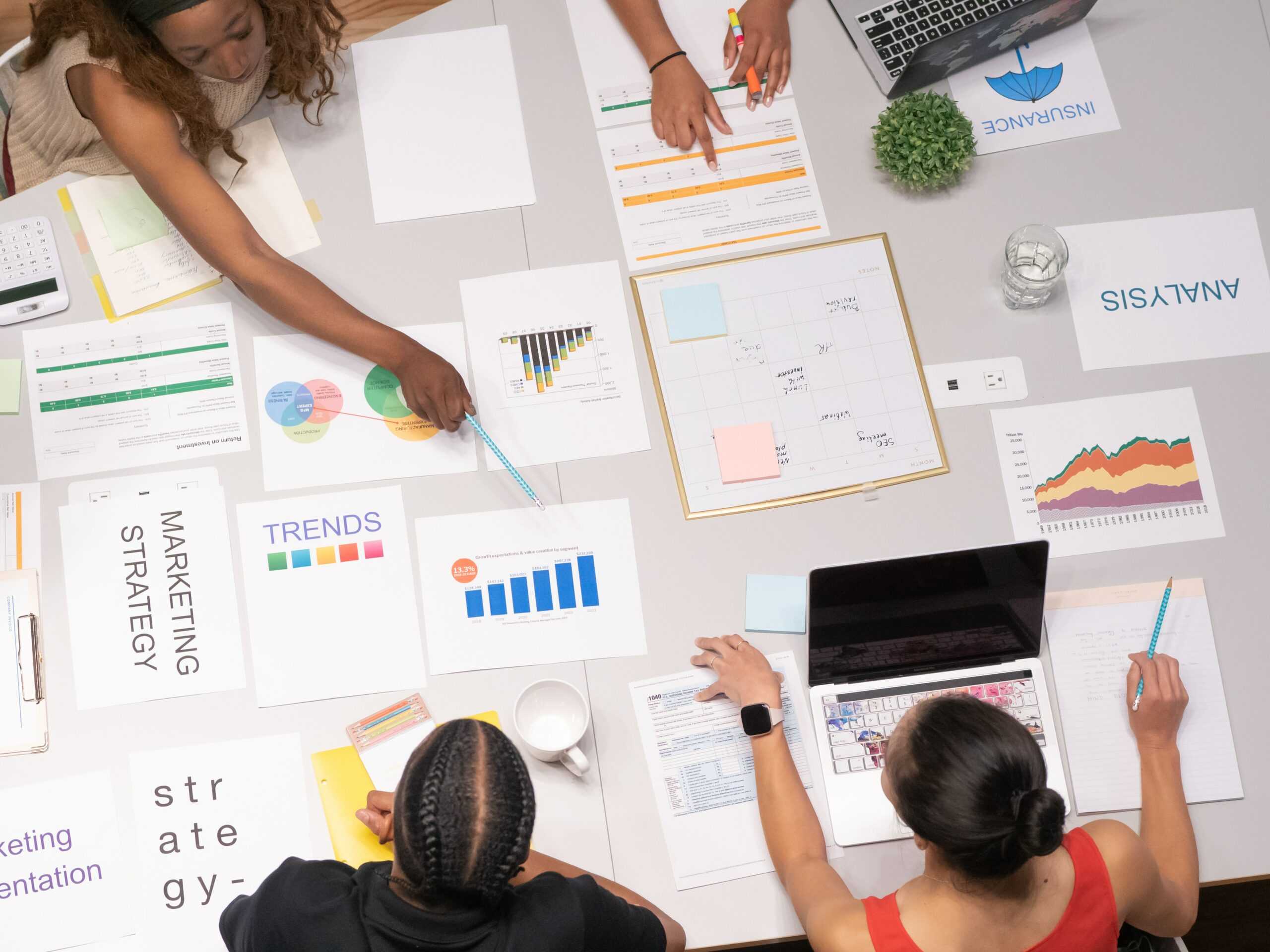 an overhead view of a large desk shows people comparing charts and graphs of their marketing work, using these tools to calculate and measure their business success